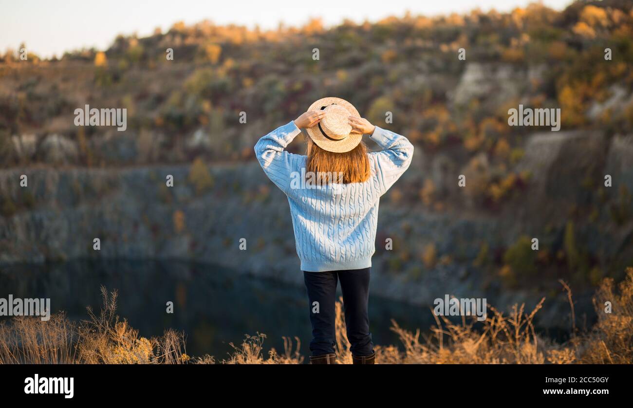 Autumn girl in blue sweater and hat standing backwards and admire nature lake view. Autumn forest colors with girl back view. Autumn portrait. Stock Photo