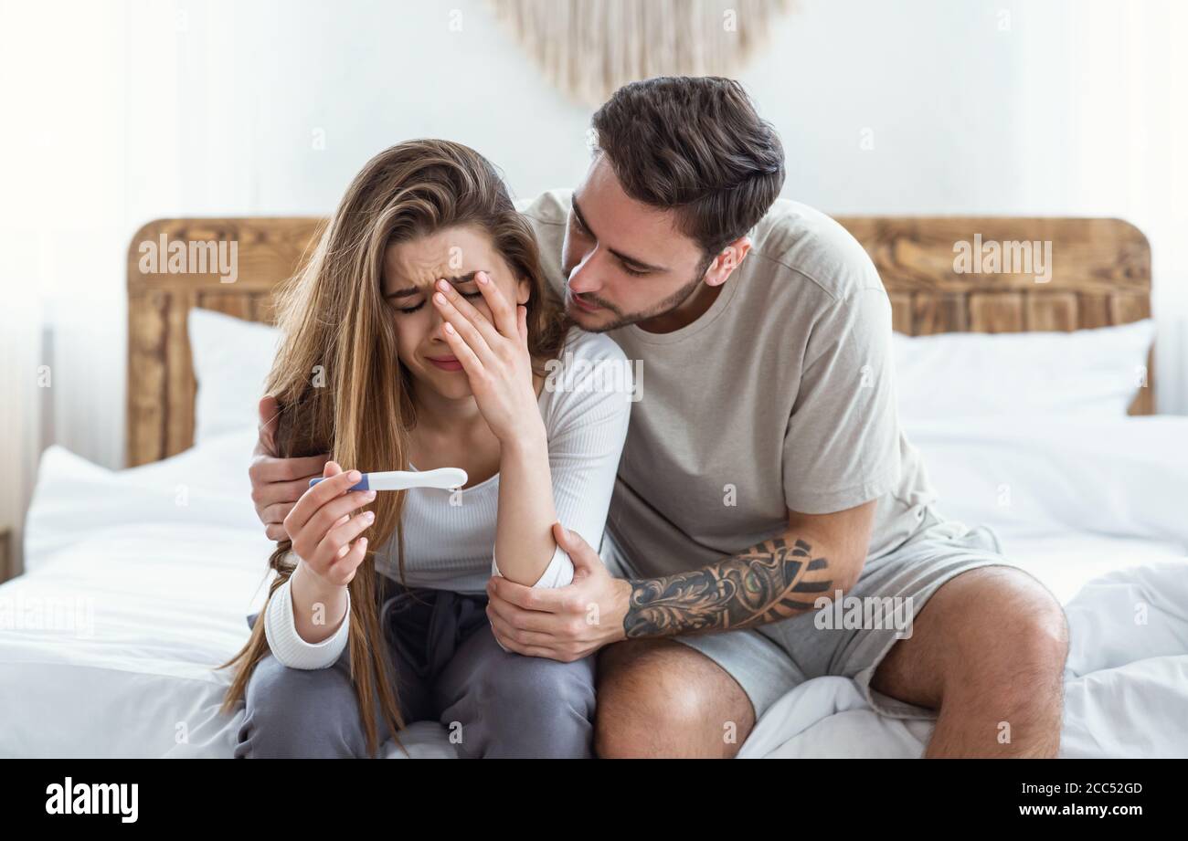 Sad man hugs crying woman and looks at pregnancy test in bedroom interior Stock Photo
