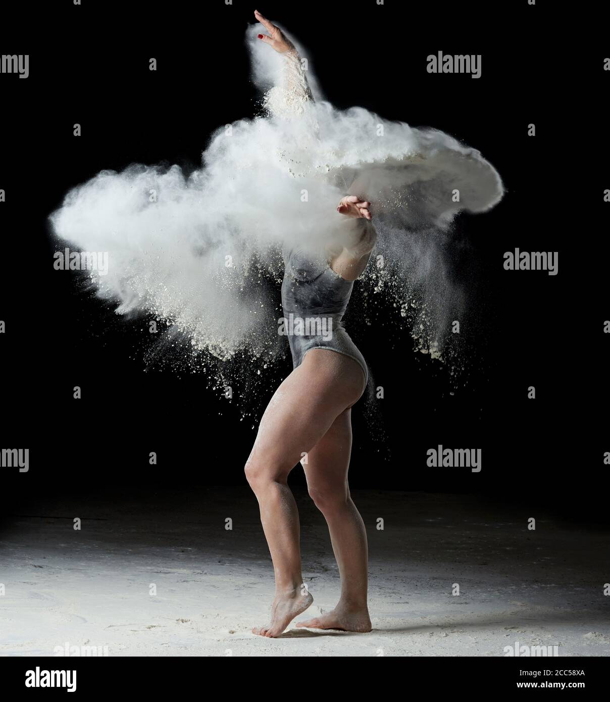 beautiful caucasian woman in a black bodysuit with a sports figure is dancing in a white cloud of flour on a black background Stock Photo