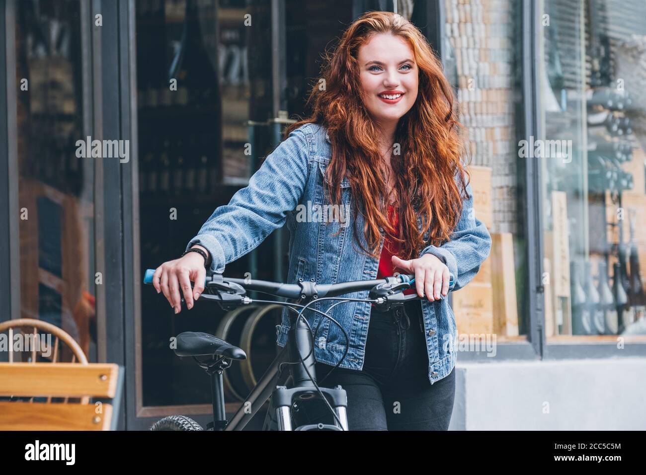 Portrait of smiling red curled long hair caucasian woman on the city street walking with a bicycle. Natural people beauty concept image. Stock Photo