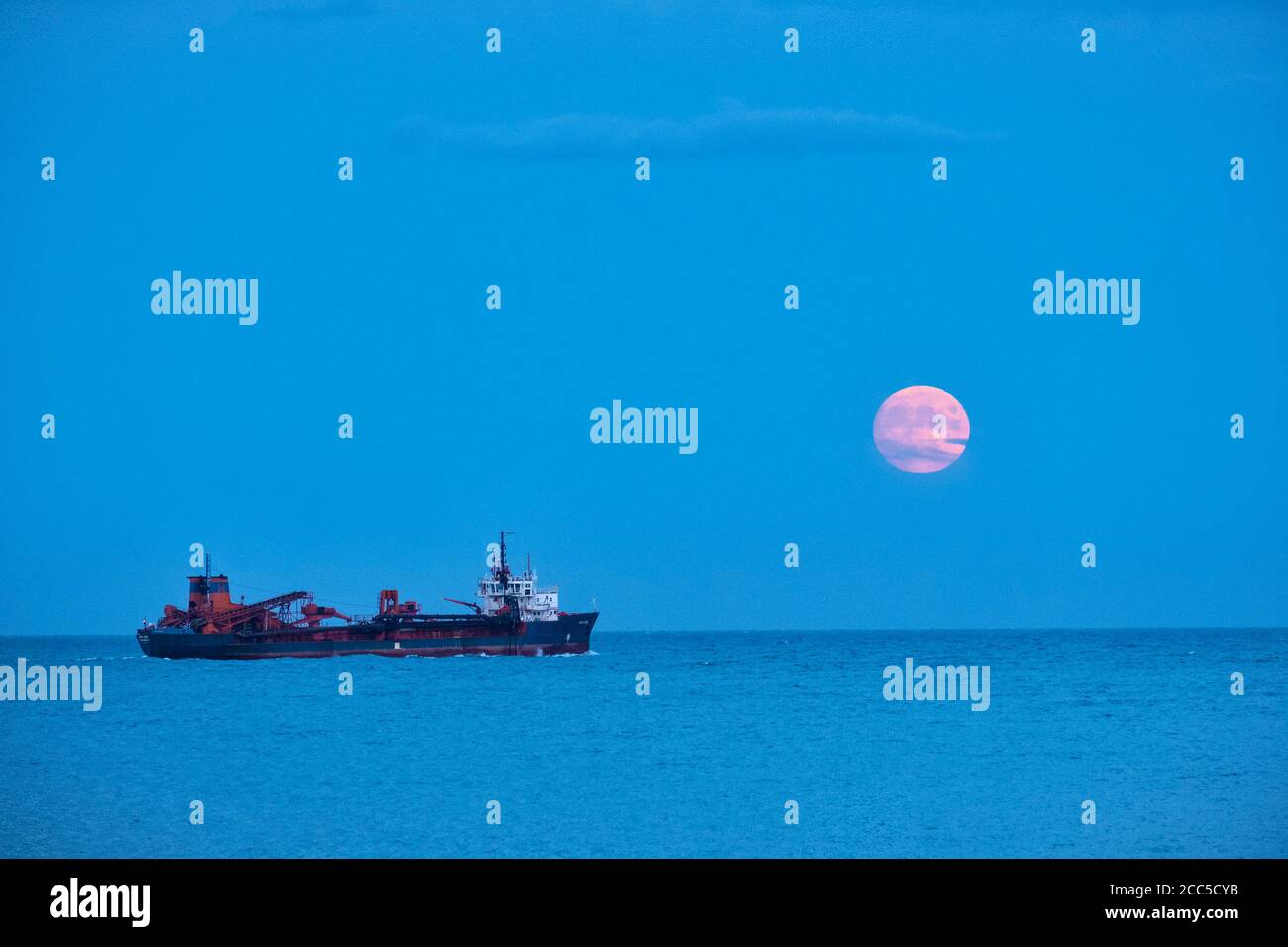 A ship (the hopper dredger Arco Avon) sailing in the English Channel off Deal, Kent, UK, under a rising full moon in August Stock Photo