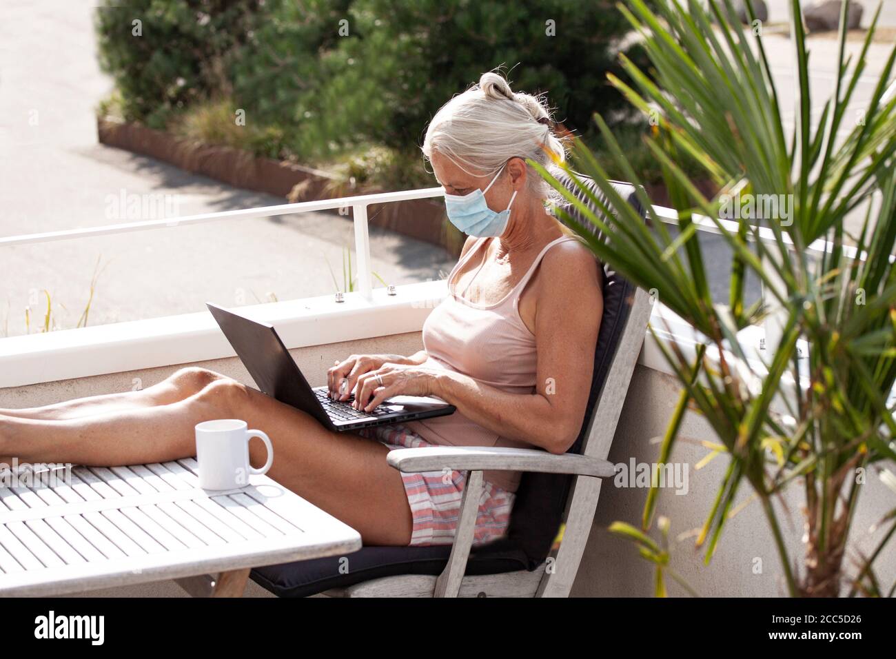 Sun tanned senior woman in shorts sitting on a balcony wearing a medical face mask to protect against Coronavirus or Covid-19 pandemic while working o Stock Photo