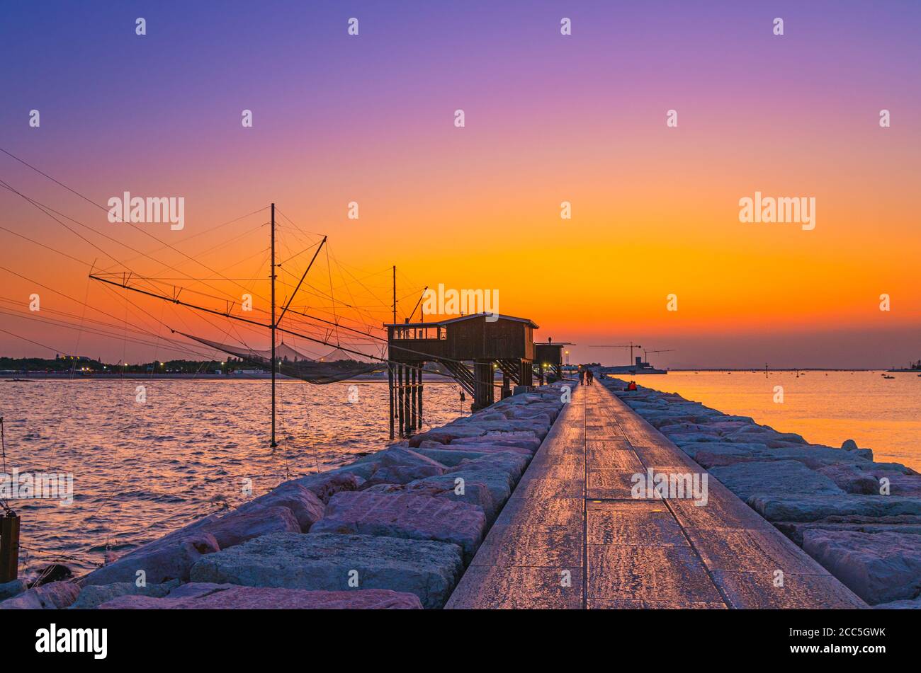 Traditional fishing station house with fishnet in water of Adriatic sea at pier Diga Sottomarina, skyline with beach, amazing yellow red sunset at twilight, dusk, evening view, Northern Italy Stock Photo