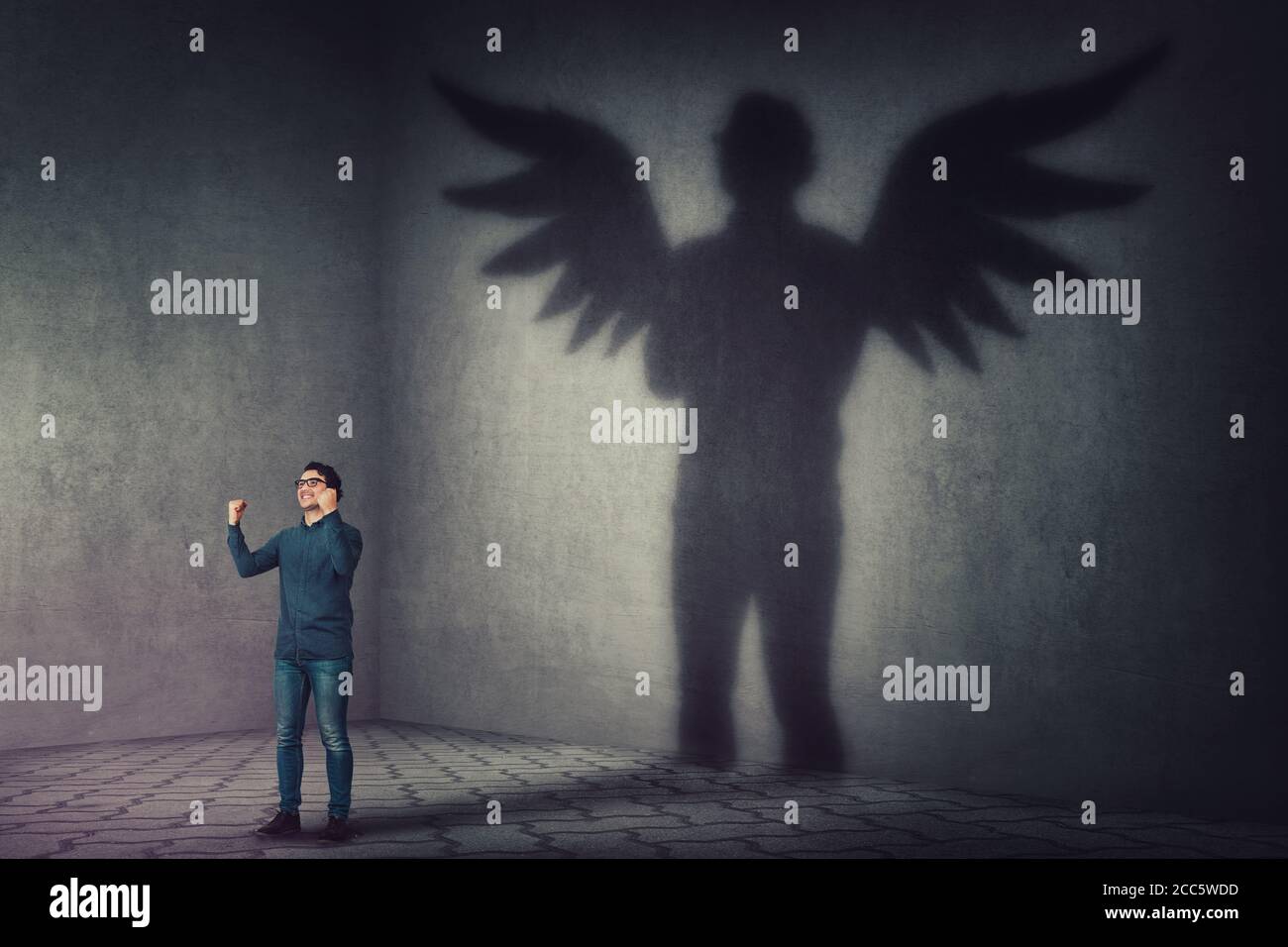 Successful businessman, raising hands up, celebrating victory, casting a superhero shadow with angel wings on a dark room wall. Self overcome, leaders Stock Photo