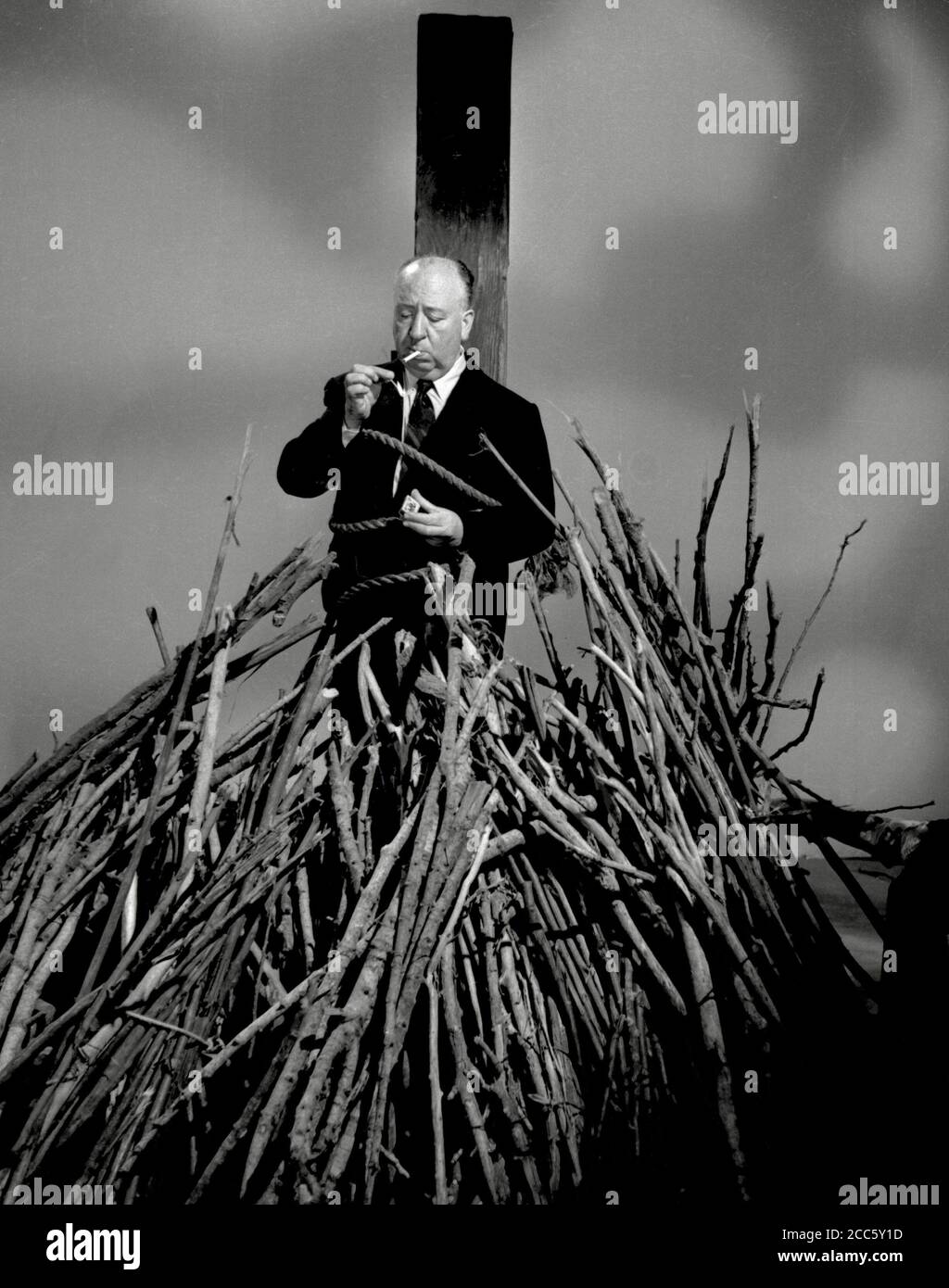 Promotional photo of film and television director Alfred Hitchcock as he lights a cigarette, while tied to a stake above a pile of sticks, for his anthology television series 'Alfred Hitchcock Presents,' August 28, 1956 CBS  File Reference # 34000-228THA Stock Photo