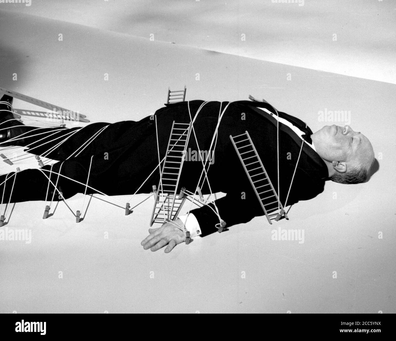Publicity photo of film and television director Alfred Hitchcock  as he lies on the floor and covered with small ladders, during his anthology television series 'Alfred Hitchcock Presents' 1962 CBS.  File Reference # 34000-284THA Stock Photo