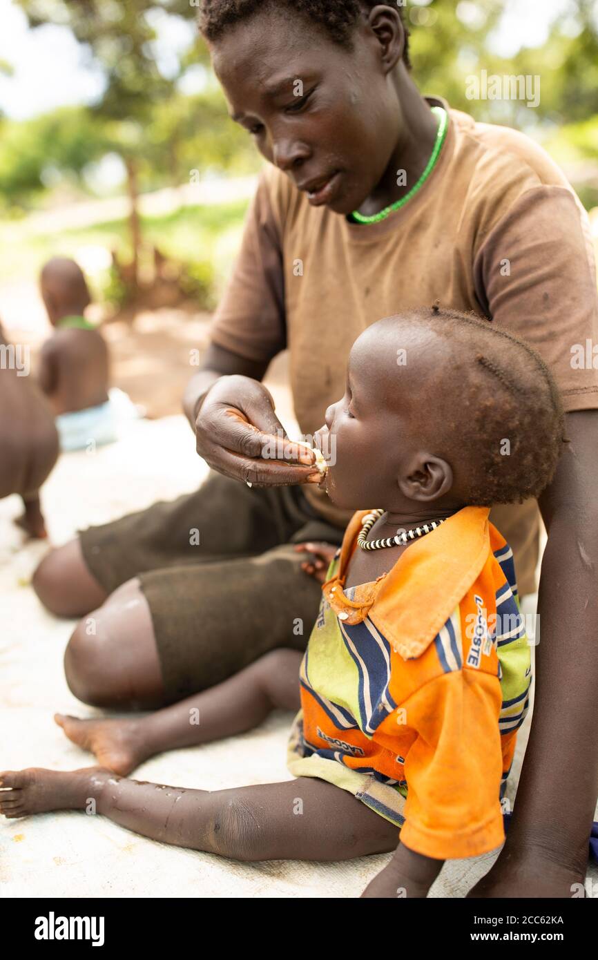 A refugee mother from neighboring South Sudan feeds her two-year-old daughter in Palabek Refugee Settlement in northern Uganda, East Africa. Stock Photo