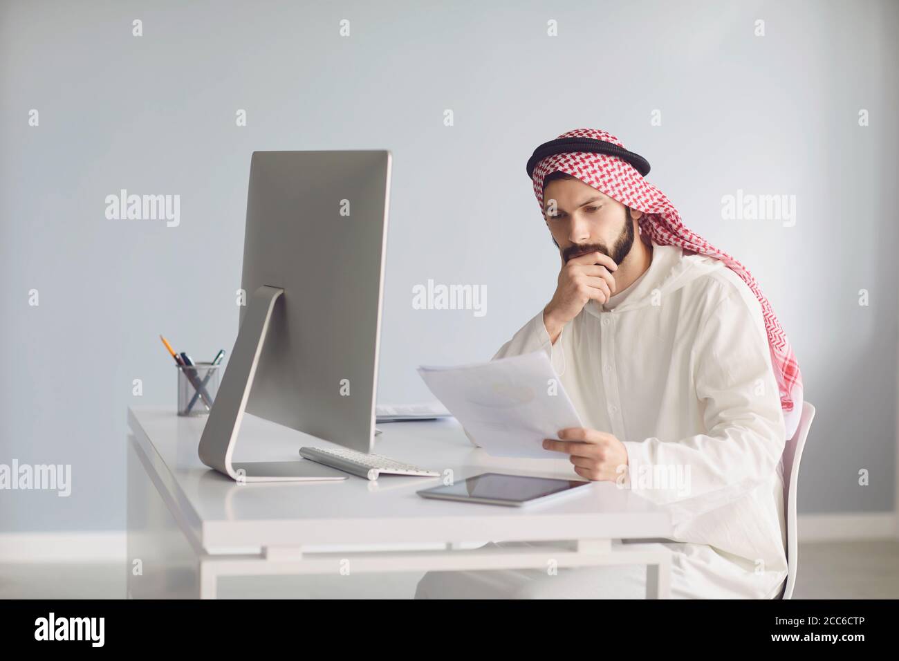 Serious Arab man thinks sitting at a table with a computer analyzes looking at paper. Stock Photo