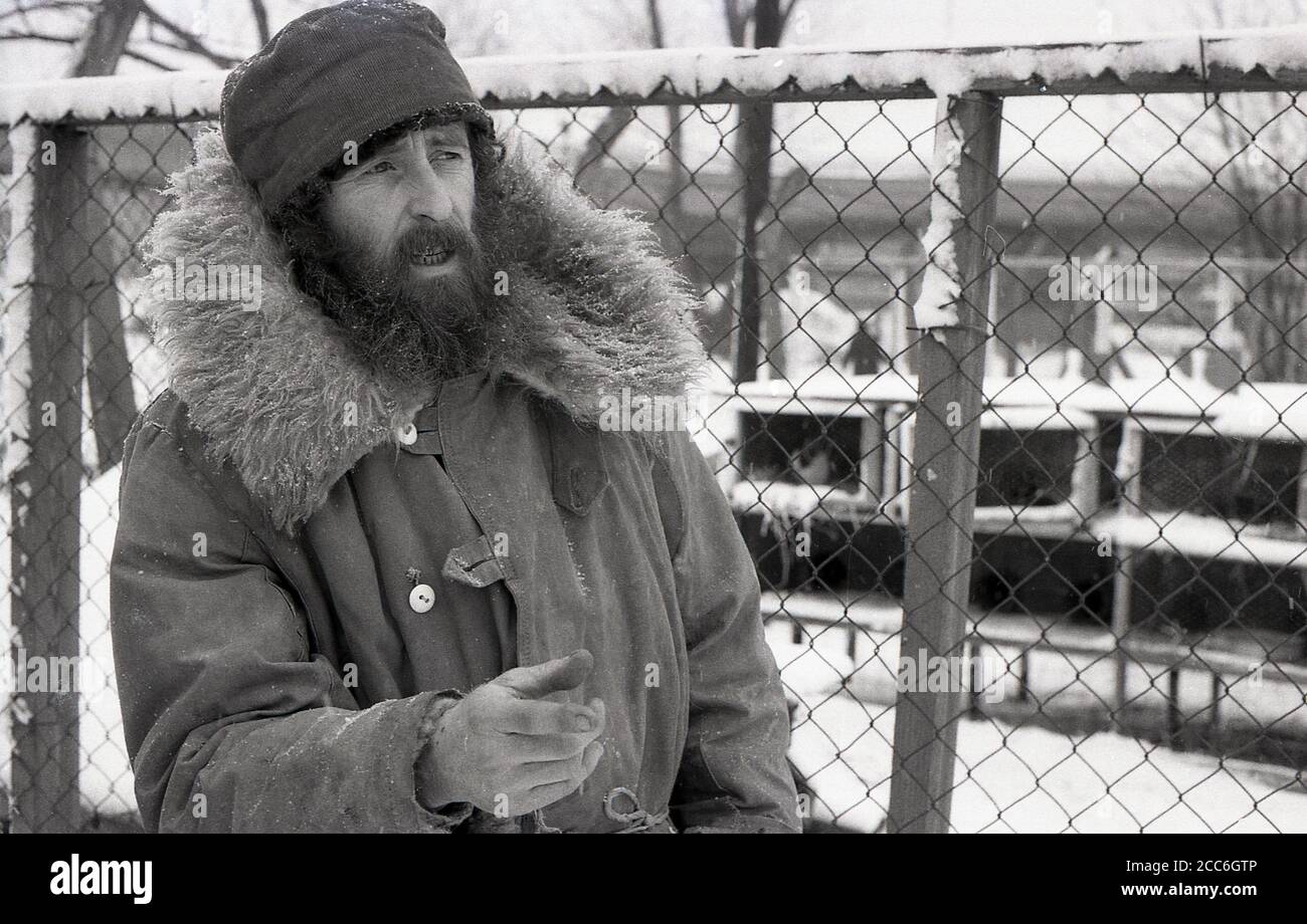1970s, historical, wintertime at a city farm inThamesmead, South-east London, snow rests on the ground in this picture of a bearded male caretaker standing at a fence outside a chicken hatch, London, England, UK. Stock Photo