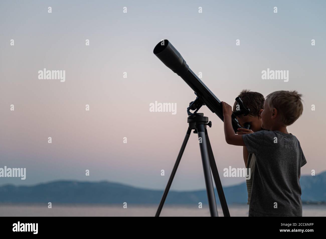 Two brothers, toddler boys, looking at the stars using a telescope by the sea on a beautiful summer night. Stock Photo