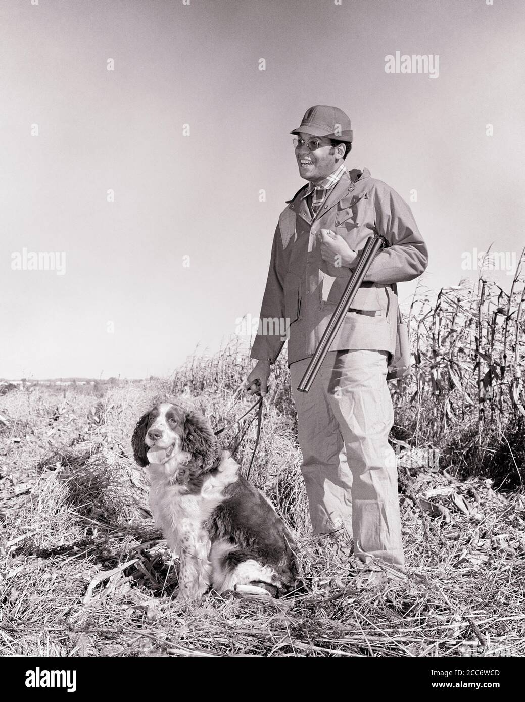 1970s SMILING AFRICAN-AMERICAN MAN UPLAND GAME HUNTER STANDING BESIDE CORNFIELD POSED WITH SHOTGUN AND SPRINGER SPANIEL DOG  - g7641 HAR001 HARS JOY LIFESTYLE SATISFACTION RURAL NATURE COPY SPACE FRIENDSHIP FULL-LENGTH PERSONS MALES HUNTER B&W HUNT ACTIVITY HAPPINESS PHYSICAL MAMMALS ADVENTURE STRENGTH AFRICAN-AMERICANS AFRICAN-AMERICAN AND CANINES RECREATION BLACK ETHNICITY FALL SEASON SPRINGER POOCH CONCEPTUAL BESIDE FLEXIBILITY MUSCLES STYLISH UPLAND CANINE CORNFIELD FIREARM FIREARMS HUNTERS MAMMAL MID-ADULT MAN POSED RELAXATION TOGETHERNESS YOUNG ADULT WOMAN AUTUMNAL BLACK AND WHITE Stock Photo