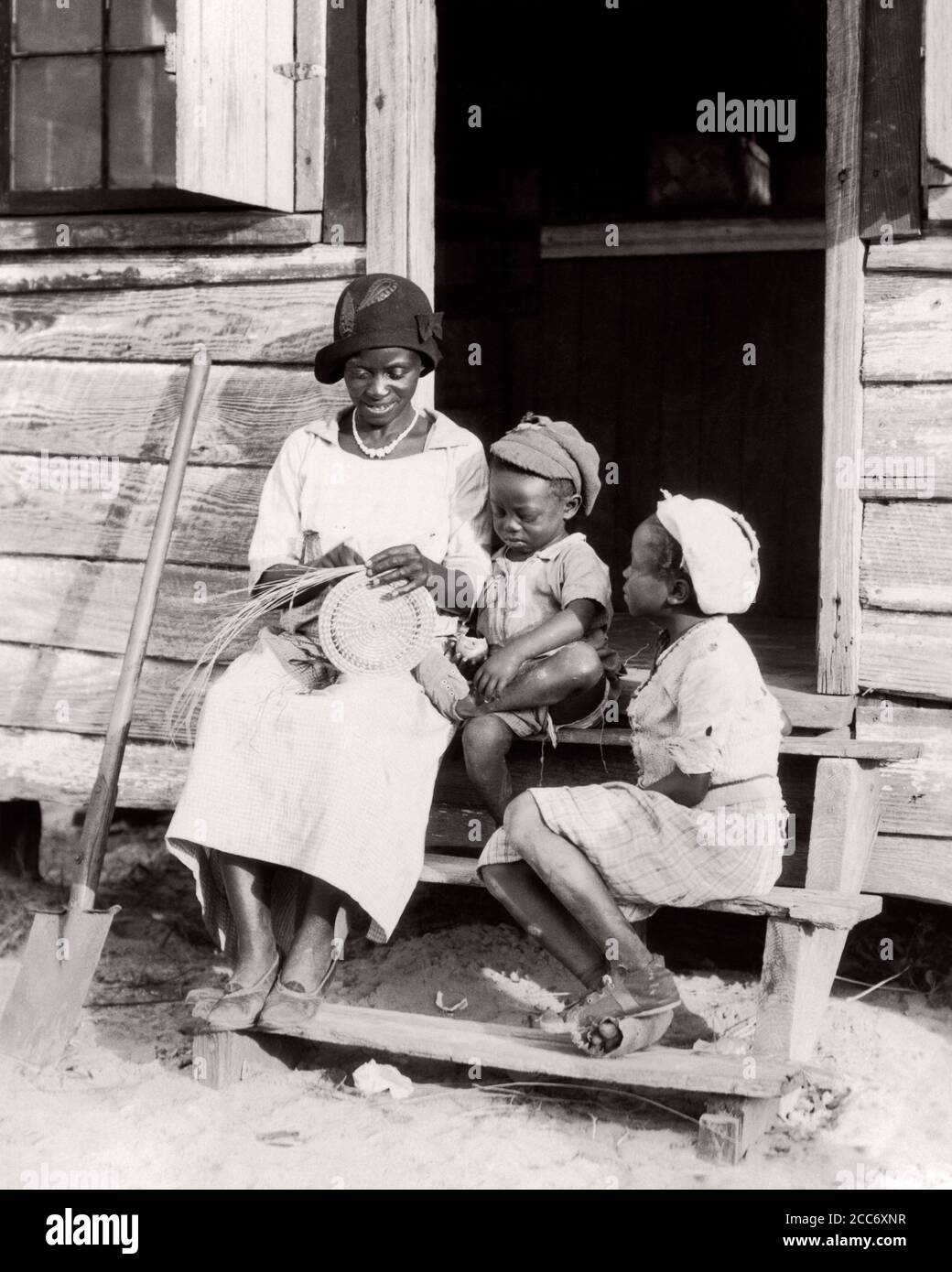 1930s AFRICAN-AMERICAN GULLAH MOTHER WEAVING A TOURIST BASKET SITTING ON STEPS TO CABIN WITH HER TWO CHILDREN SOUTH CAROLINA USA - n1209 HAR001 HARS 3 ART TRAVEL MOM CLOTHING STRESS NOSTALGIC PAIR BEAUTY COMMUNITY STEPS HER MOTHERS OLD TIME BUSY NOSTALGIA BROTHER OLD FASHION SISTER POVERTY 1 JUVENILE STYLE LOST SONS FAMILIES LIFESTYLE FEMALES HOUSES BROTHERS POOR RURAL HOME LIFE UNITED STATES COPY SPACE FULL-LENGTH LADIES DAUGHTERS PERSONS RESIDENTIAL UNITED STATES OF AMERICA CARING MALES BUILDINGS CABIN CRAFT SIBLINGS SISTERS B&W SADNESS NORTH AMERICA FREEDOM NORTH AMERICAN SKILL SOUVENIR Stock Photo