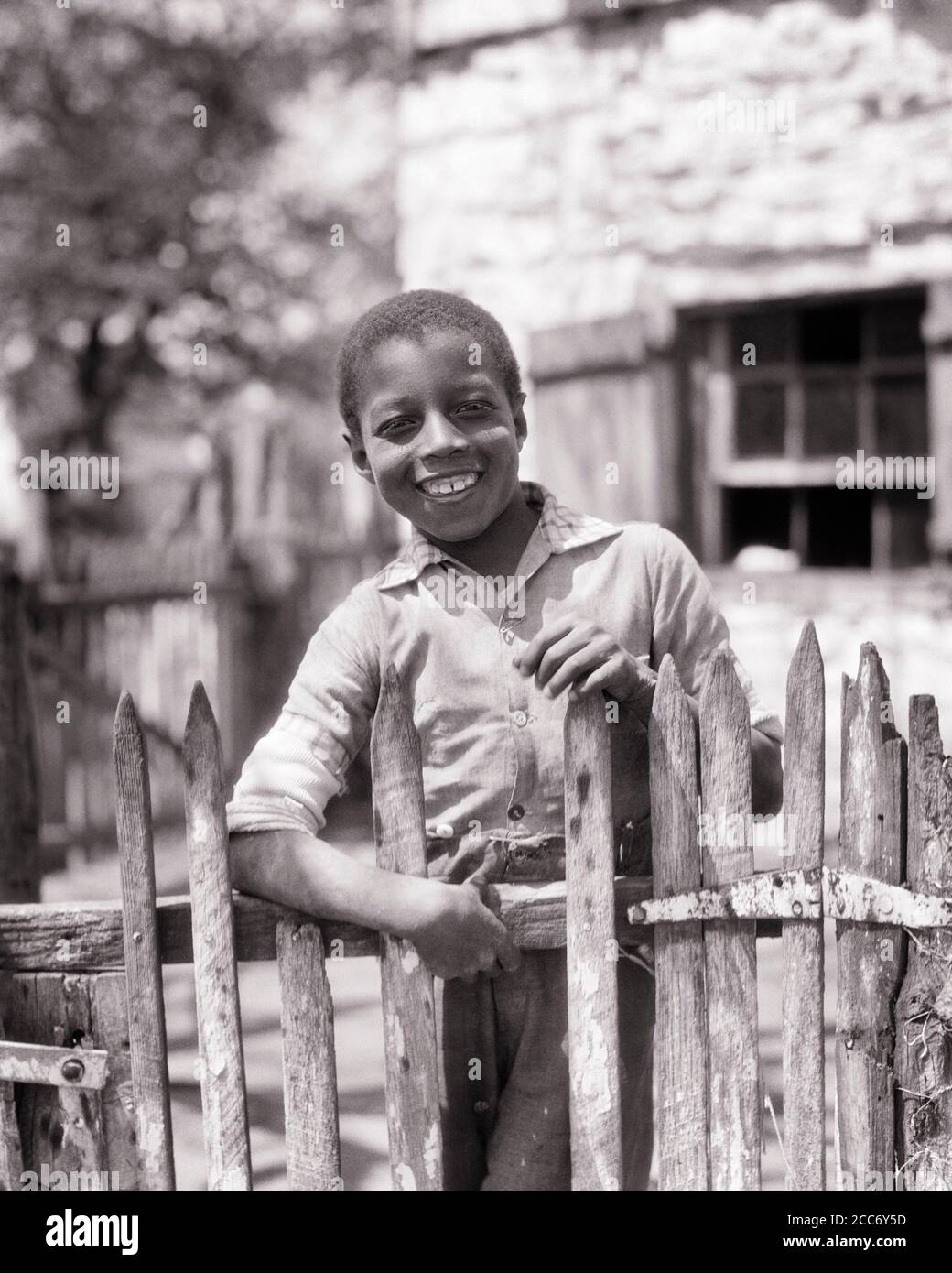 1930s SMILING ANONYMOUS RURAL AFRICAN-AMERICAN BOY STANDING LOOKING AT CAMERA AT HOME BEHIND PICKET FENCE - n936 HAR001 HARS MALES EXPRESSIONS B&W EYE CONTACT CHEERFUL AFRICAN-AMERICANS AFRICAN-AMERICAN BLACK ETHNICITY PRIDE AT SMILES CONCEPTUAL JOYFUL DISADVANTAGED ANONYMOUS IMPOVERISHED JUVENILES BLACK AND WHITE HAR001 OLD FASHIONED AFRICAN AMERICANS Stock Photo