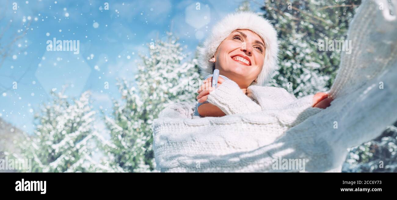 Happy smiling woman portrait in knitwear and fur hat enjoys with a hot cup of tea on sunny winter day during snowy forest walk. Stock Photo