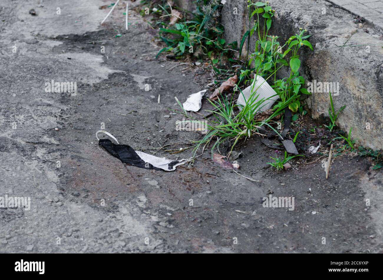 homemade protection mask thrown on the street beside other garbage accumulating dirt on the curb. Stock Photo