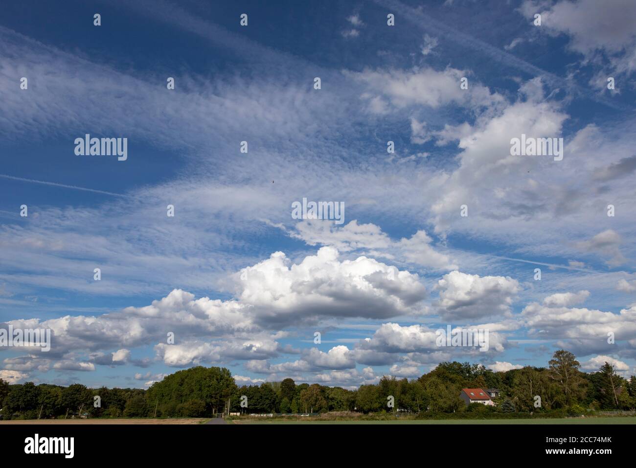blue sky with clouds and condensation trails above a small village and trees, outdoors Stock Photo