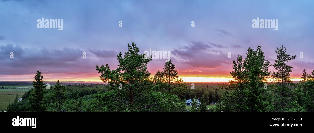 Aerial panorama of midnight sun above light green northern scandinavian forest - fire burning clouds, fir tree tops, cold summer night, yellow red col Stock Photo
