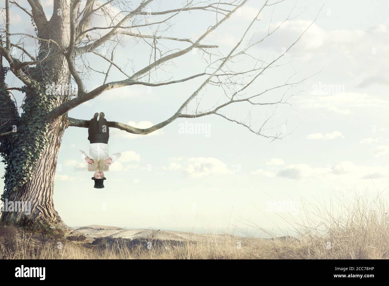 surreal moment of man reading a book hanging from a branch on  head down Stock Photo
