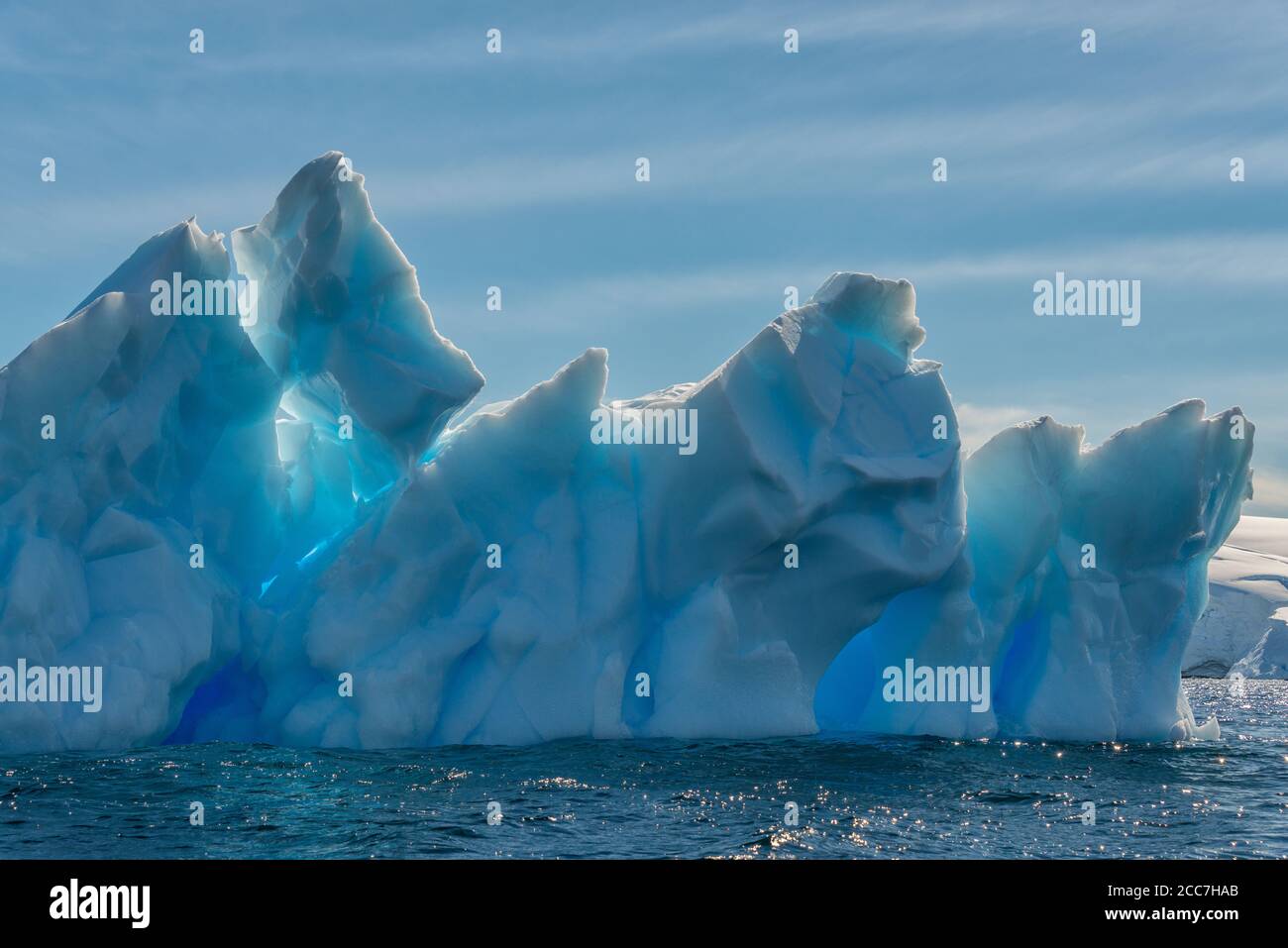 An iceberg with a range of blue hues floats in the sea near the glacier covered coast on a sunny afternoon in Antarctica. Stock Photo