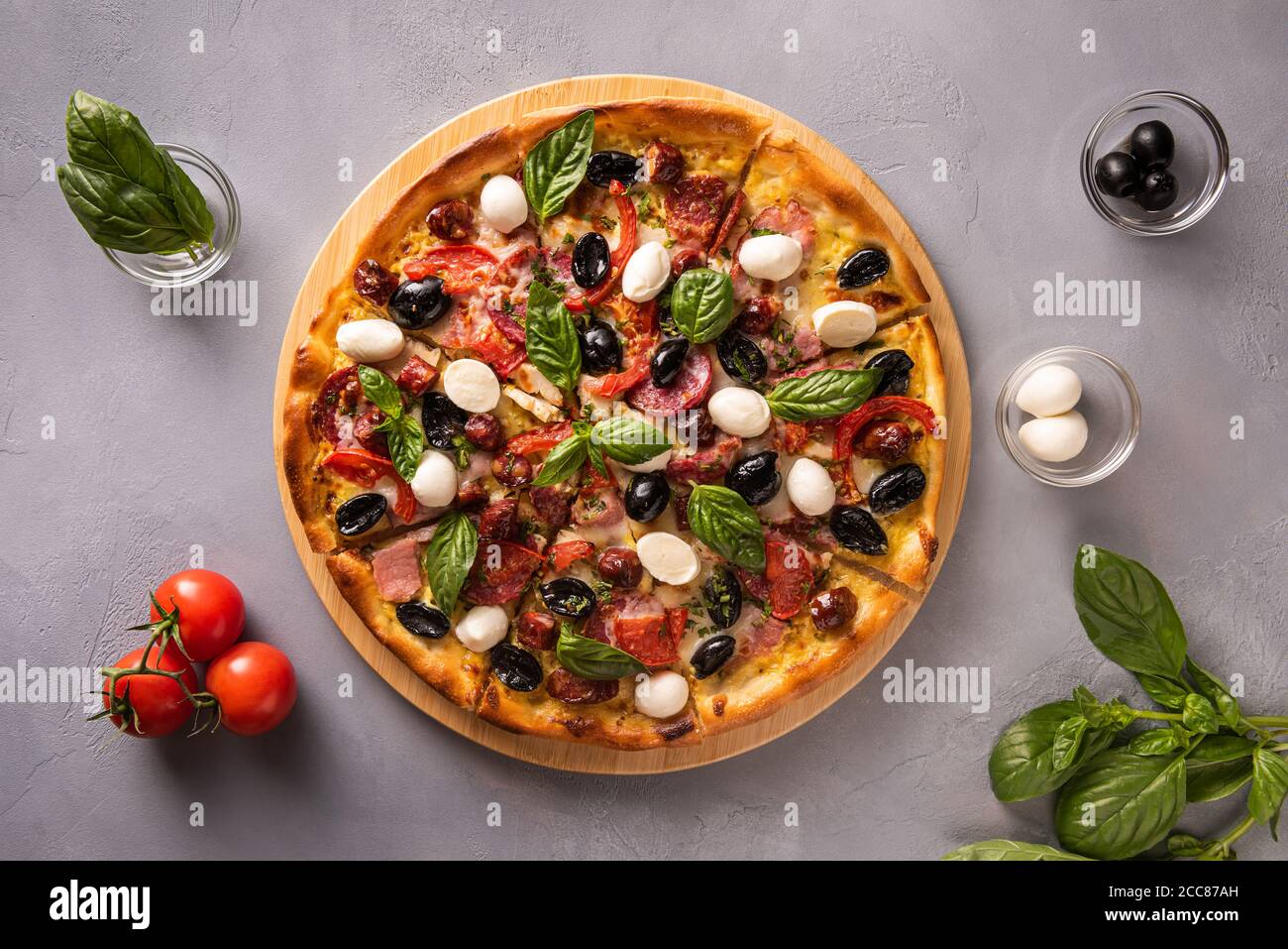 Italian pizza with ham, salami, black olives, mozzarella cheese, red tomatoes and basil leaves on gray textured stone background and few ingredients i Stock Photo