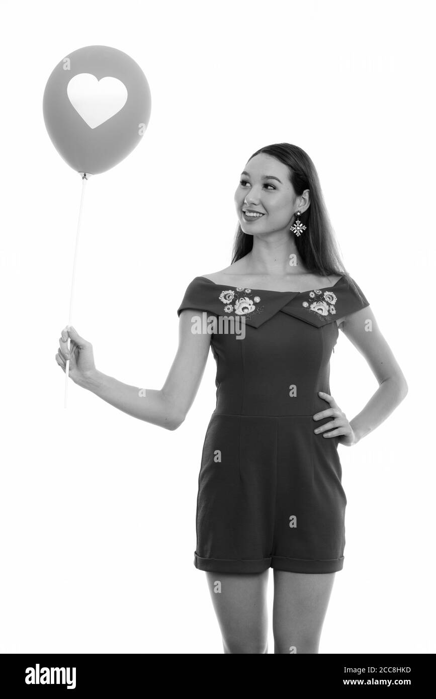 Studio shot of young happy beautiful Asian woman smiling and standing while looking at balloon with heart sign Stock Photo