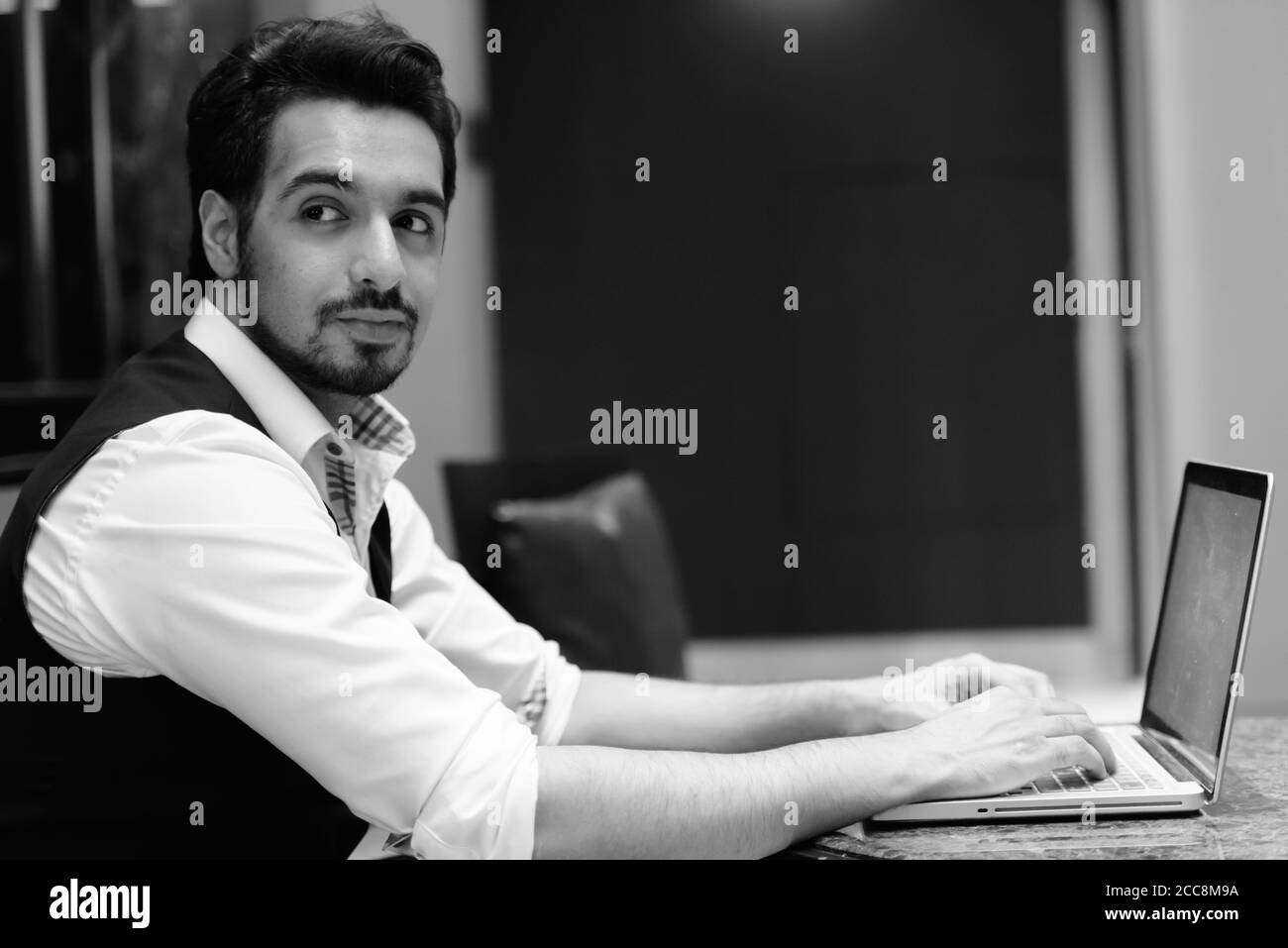 Young handsome Indian man using laptop while looking back in the lobby of hotel Stock Photo