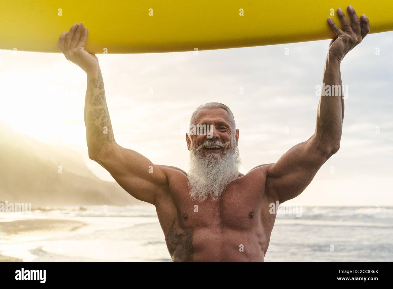 Happy fit senior having fun surfing at sunset time - Sporty bearded man training with surfboard on the beach Stock Photo