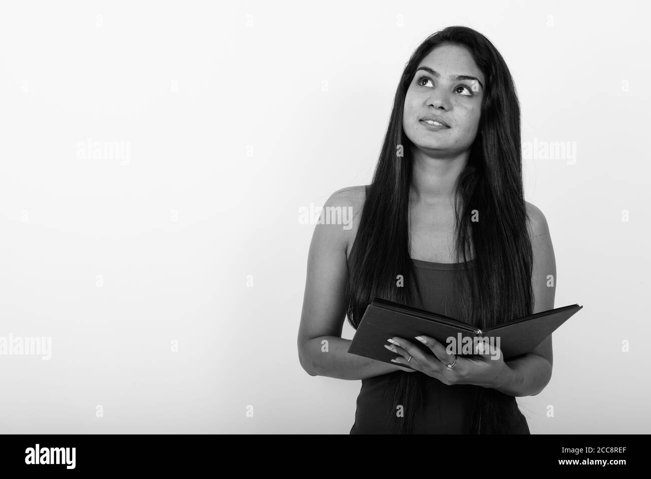 Studio shot of young happy Indian woman smiling and thinking while holding open book against white background Stock Photo