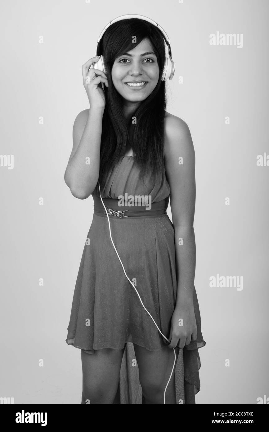 Studio shot of young happy Persian woman smiling and standing while listening to music against gray background Stock Photo