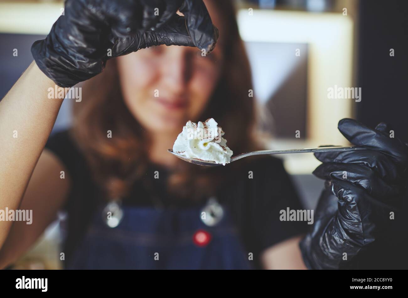 Barista wearing medical latex black gloves, hold spoon with whipped cream. Process of making dessert in cafe.Blurred image, selective focus Stock Photo