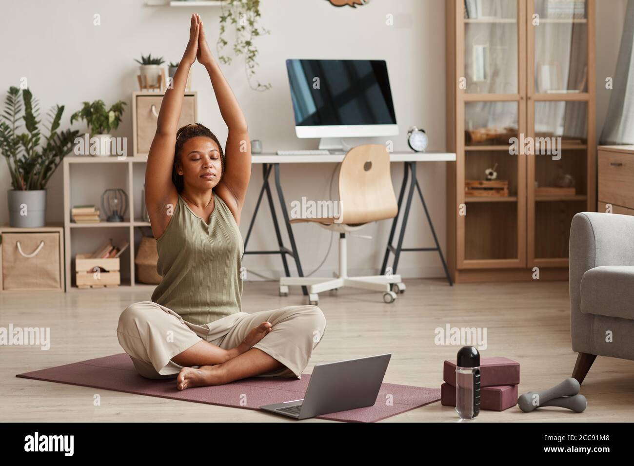 Young woman sitting on exercise mat in front of laptop and doing yoga ...