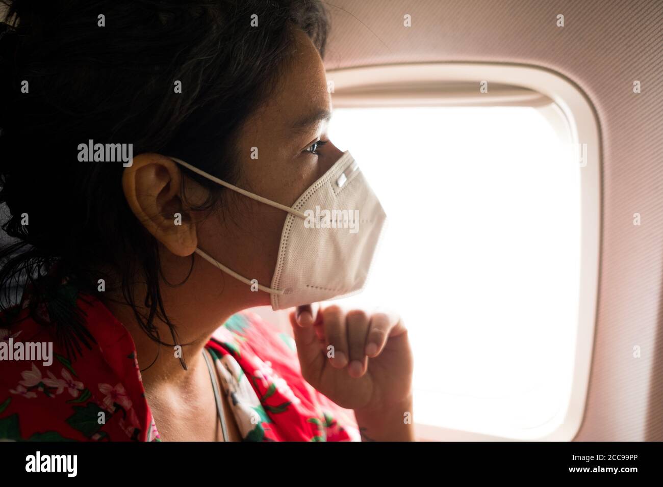 worried young woman inside plane wearing protective face mask, going on vacation and traveling during the coronavirus pandemic, which has caused a cri Stock Photo