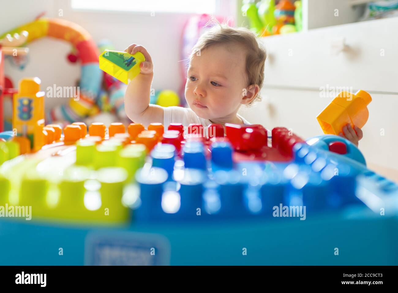 Baby girl (16 months) sitting on the floor and playing in her bedroom with colourful Mega Bloks® plastic blocks near her white wooden chest of drawers Stock Photo