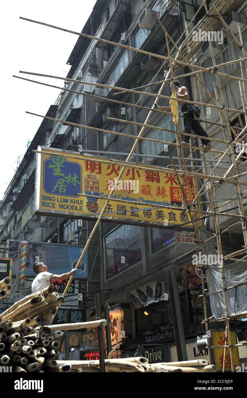 Two men standing in scaffolding made from bamboo poles, one passing a long bamboo pole up to the other. Kowloon, Hong Kong, China Stock Photo