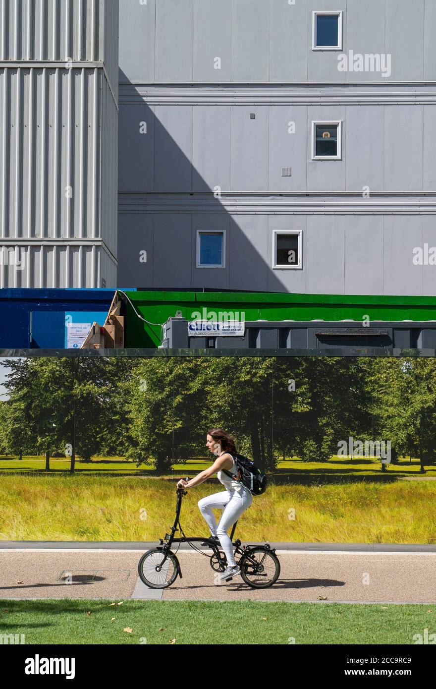 A cyclist rides past a hoarding showing a green woodland scene, outside a building site in north London, as people enjoy the warm weather after recent heavy rainfall. Stock Photo