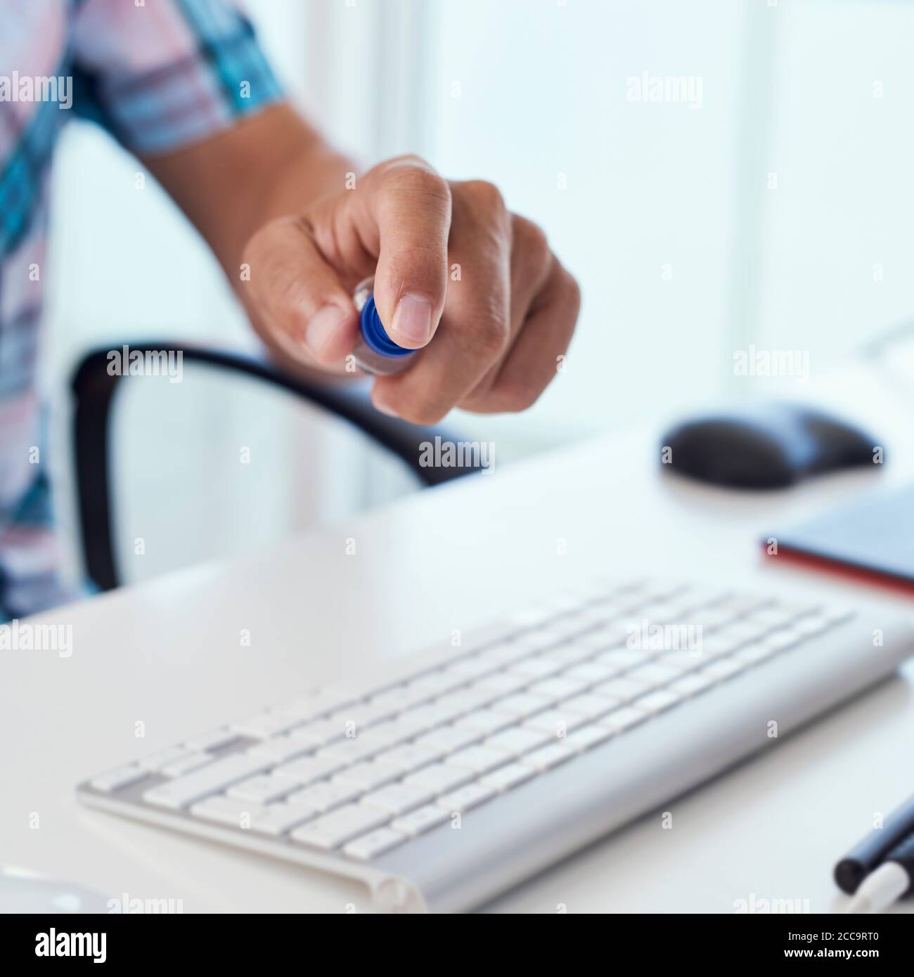 closeup of a young caucasian man sitting at his office desk disinfecting his computer keyboard by spraying a disinfectant from a bottle Stock Photo