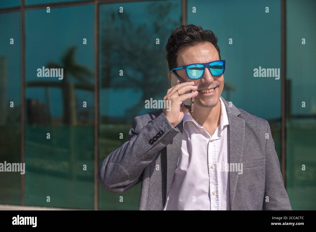 Happy adult male entrepreneur in smart casual clothes and sunglasses smiling and looking away while answering phone call on city street Stock Photo