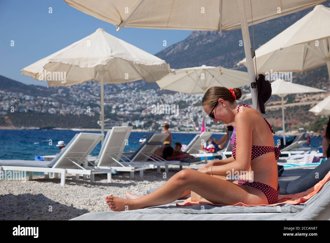 A young woman  ( model released ) sitting on sunlounger at the Kalkan beach club,  Kishla, with the town of Kalkan, Turkey, in the background. Kalkan Stock Photo