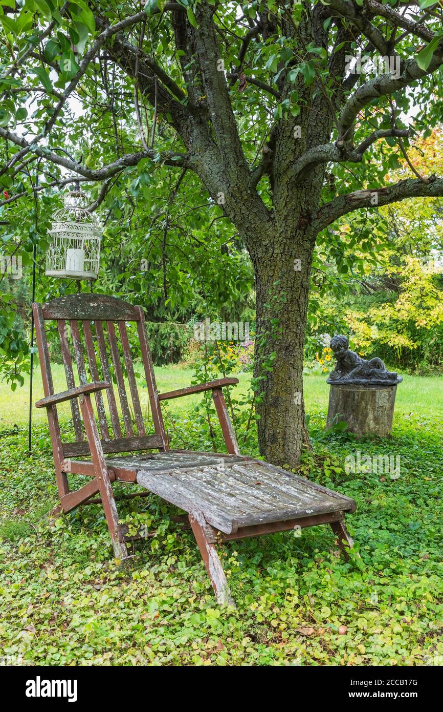 Old wooden teak wood lounge chair with lichen growth and statue of young boy on tree stump observing a snail under Pyrus - Pear tree. Stock Photo