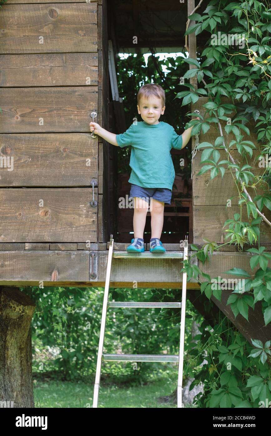 Small boy stands on stairs on treehouse in green garden. Summer time Stock Photo