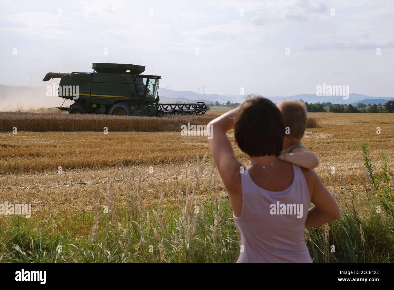 Harvesting wheat in an autumn field. Woman and child look at the working of a modern green combine Stock Photo