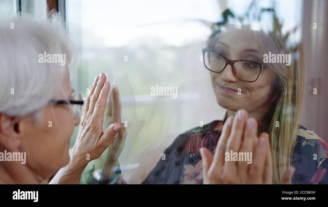 Young lady in quarantine speaking with her grandmother through the glass window. Protecting vulnerable from covid infection. High quality photo Stock Photo