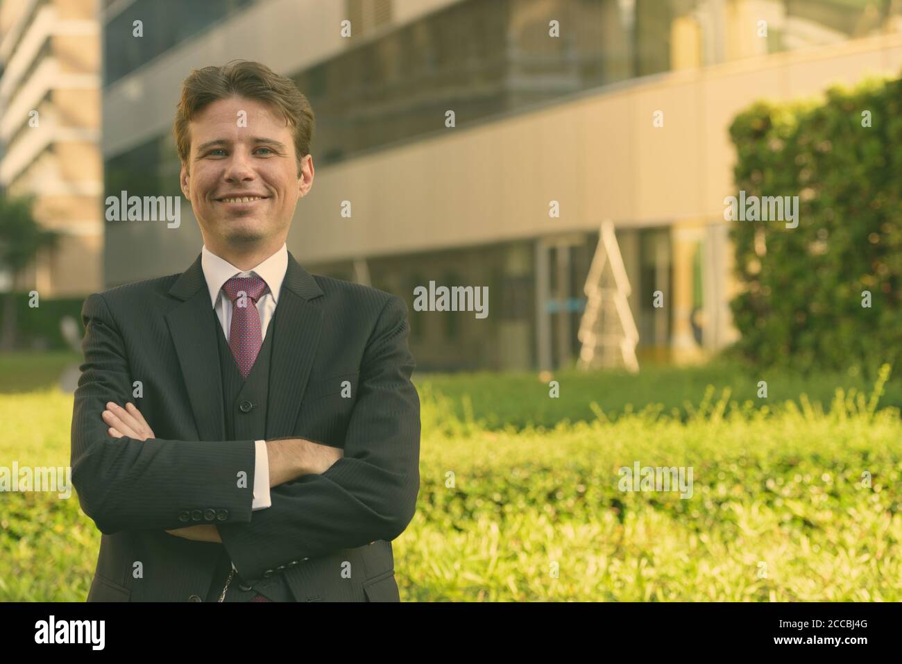 Happy businessman smiling with arms crossed in front of modern garden ...