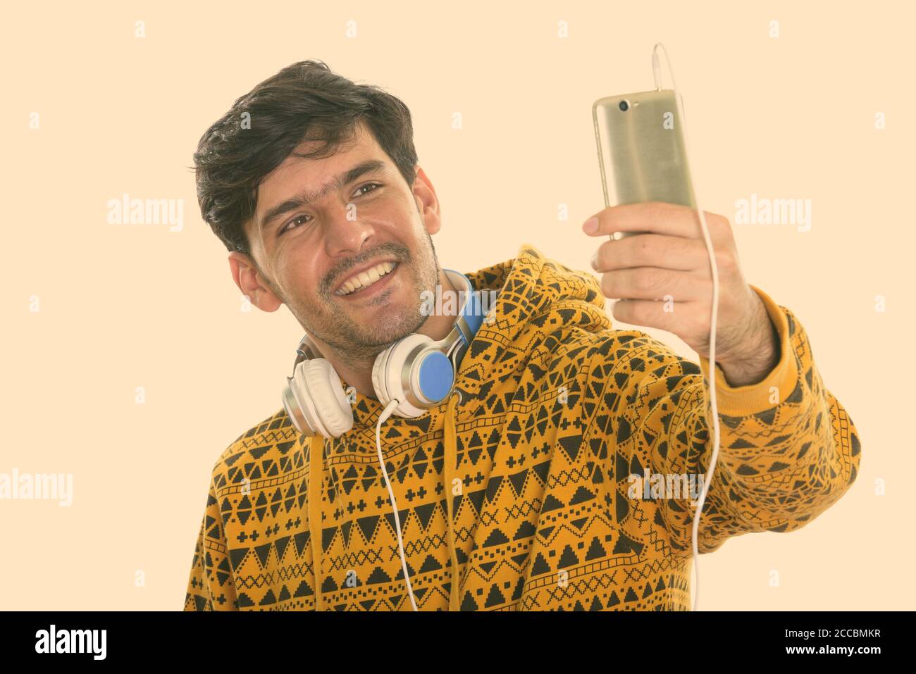 Young happy Persian man smiling while wearing headphones around neck and taking selfie picture with mobile phone Stock Photo