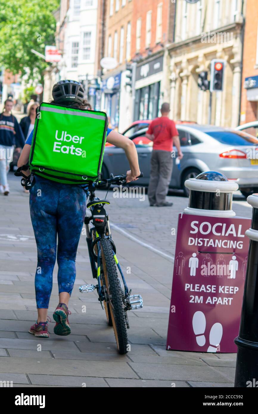 Winchester, United Kingdom - 16 July 2020: Uber Eats bike courier walking on pavement by the street, passing by social distancing information banner Stock Photo