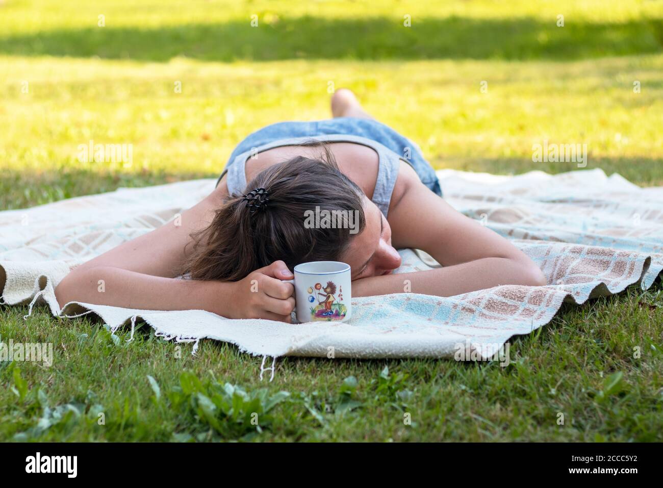 young beautiful woman lying on her stomach on a blanket and holding a cup in her hand, on the grass, sunny summer day Stock Photo