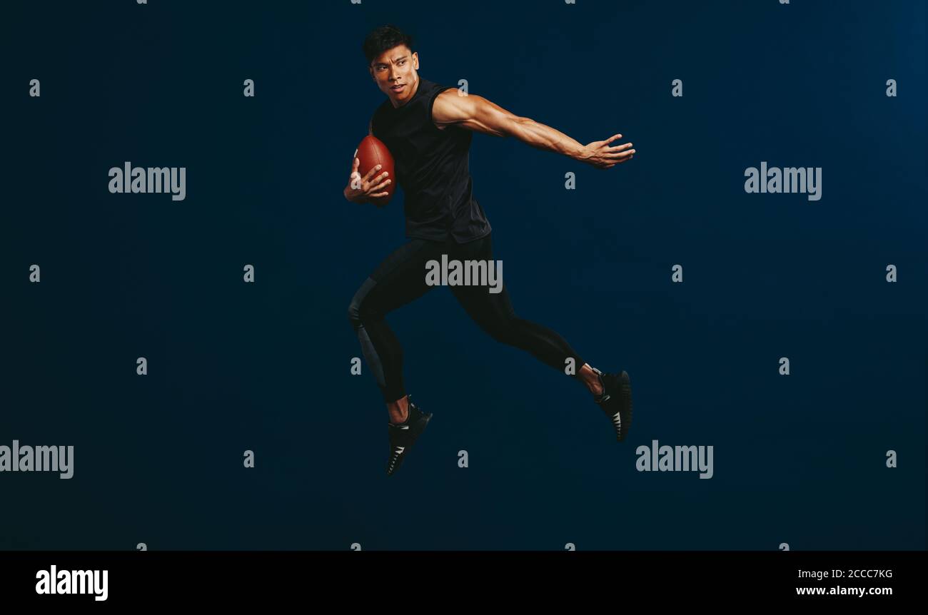 American football player holding ball and jumping against dark background. Sportsman practising football. Stock Photo