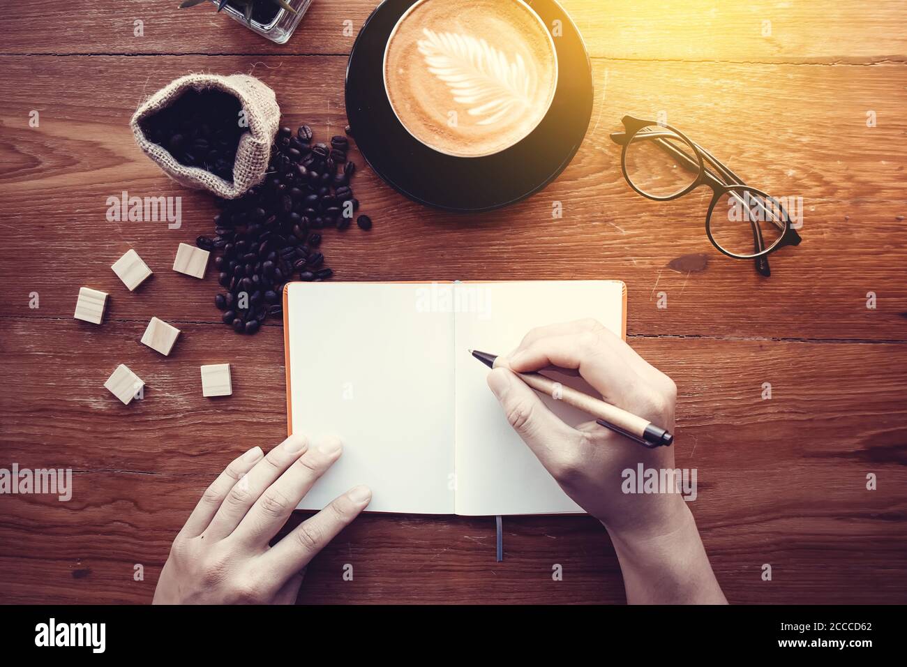 Education background concept : close up man with pencil in hand writing in notebook on wood table with coffee cup, top view. Vintage light effect. Stock Photo