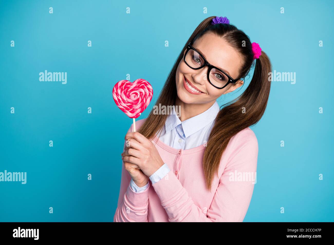 Portrait of positive cheerful college student girl hold heart shape lollipop stick enjoy lesson break wear good look clothes isolated over blue color Stock Photo