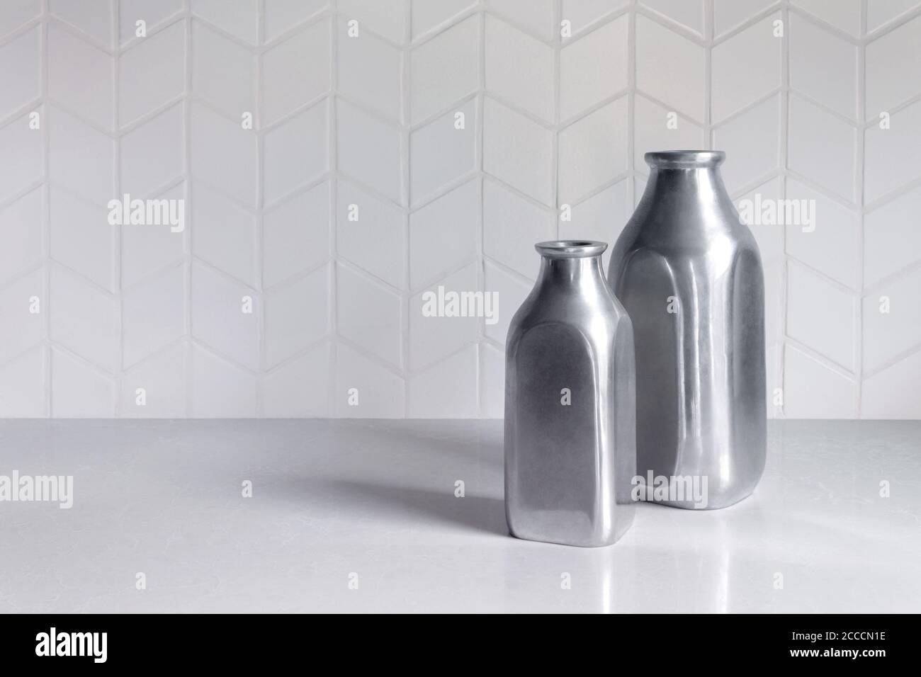 two empty silver colored metal bottles or vases on a white counter with a white chevron tile pattern backsplash with copy space Stock Photo